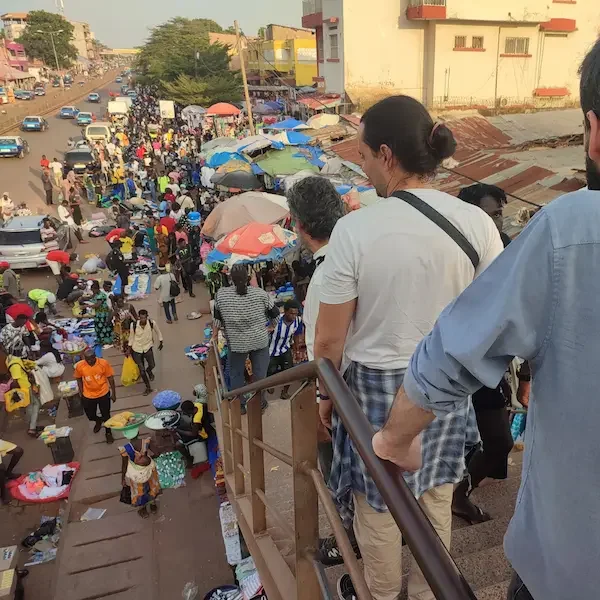 Mercado de Bissau Personas bajando unas escaleras junto a un mercado concurrido con puestos y sombrillas.