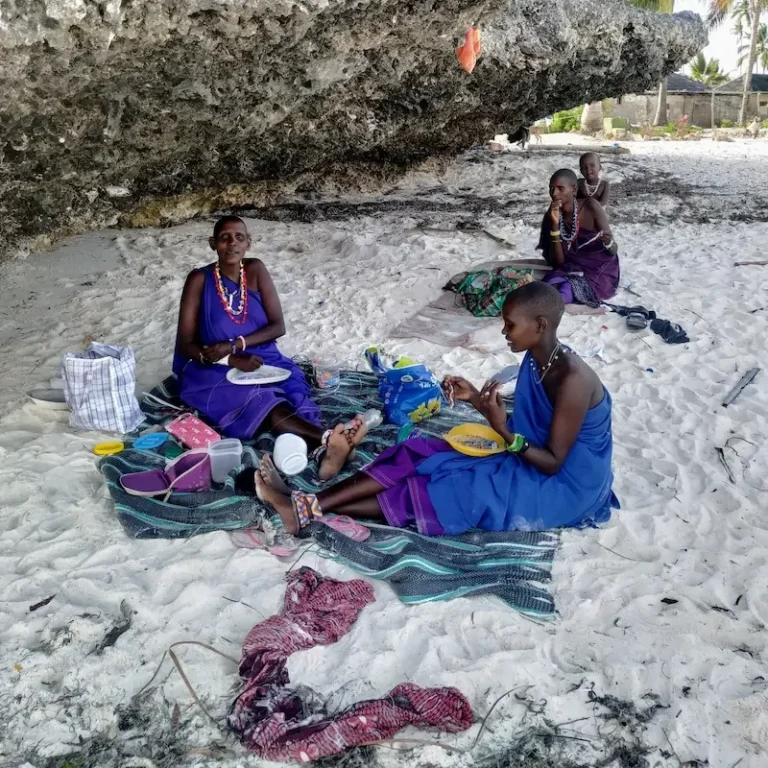 Conexión humana Tres mujeres massai sentadas sobre telas en la arena bajo una gran roca, con una mujer y un niño al fondo.