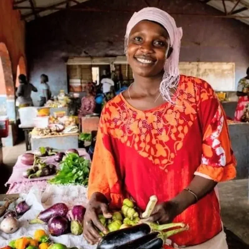Mercado en Gambia Mujer sonriente detrás de un puesto con berenjenas y otras verduras en un mercado.